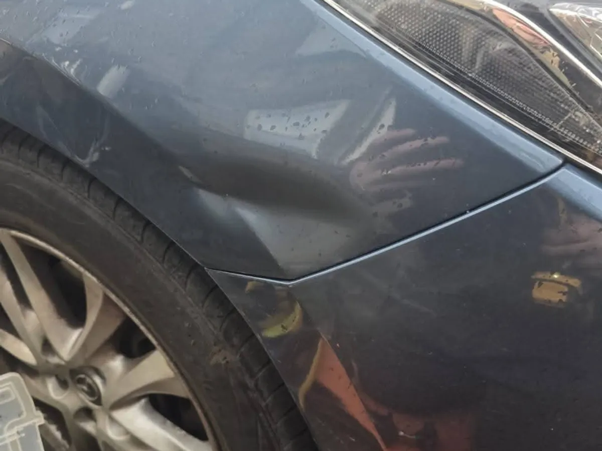 The front wing of a dark blue Mazda in the wet, the reflection breaking up over damage above the wheel arch.