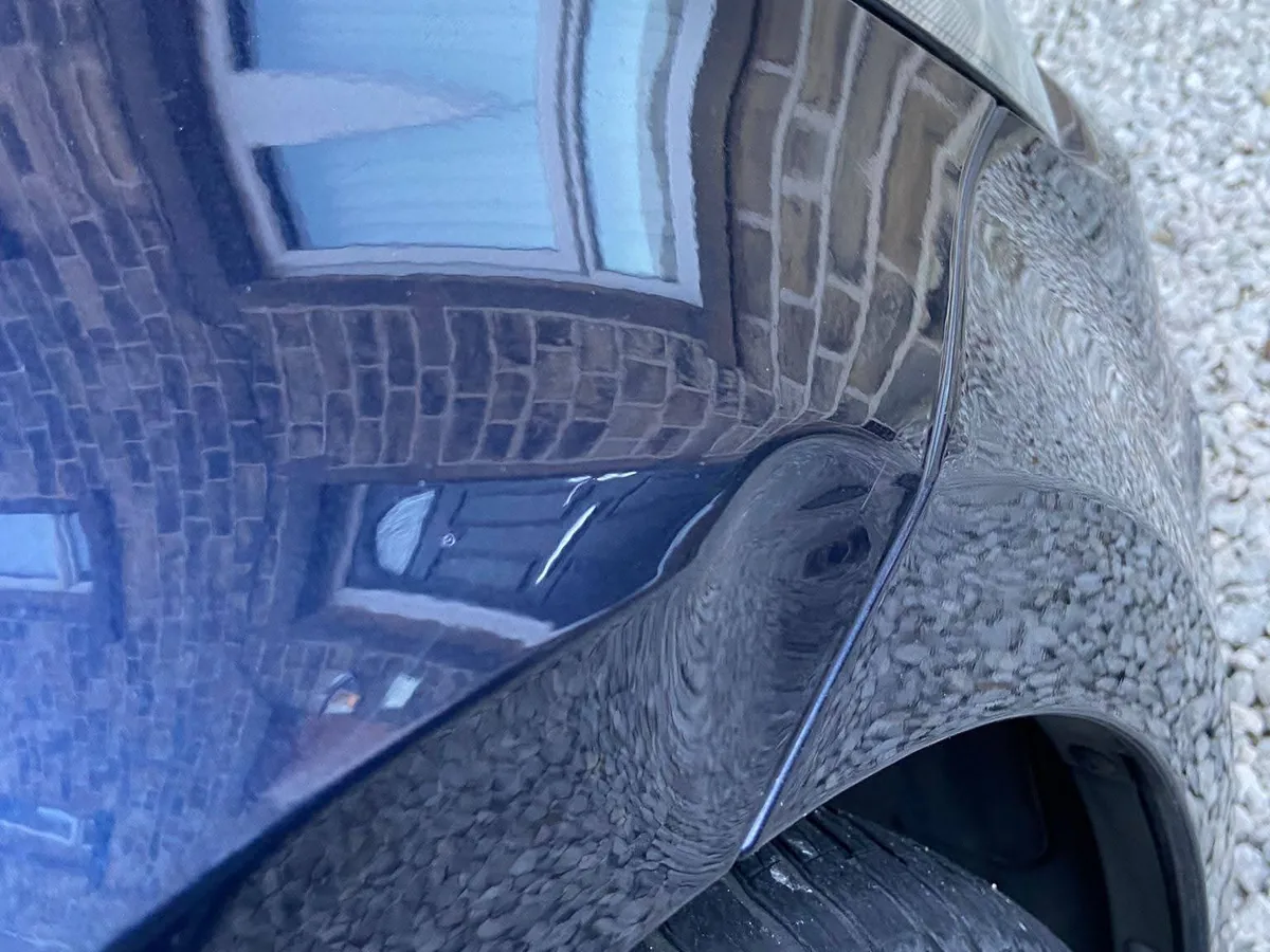 A dark blue rear quarter panel with a brick house reflected in it, the reflection bowing where the panel is dented above the wheel arch.