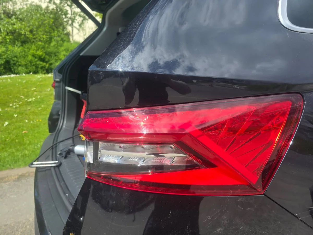 The same black quarter panel after repair, photographed with the boot open, the hedge behind now reflecting evenly across the paint.